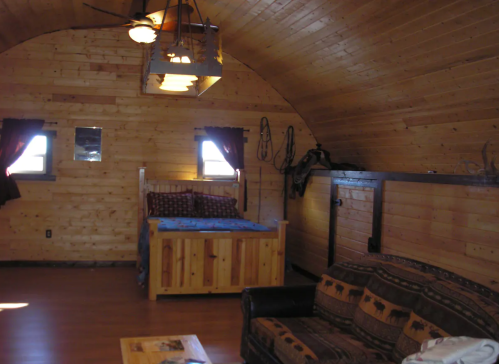 Cozy wooden interior of a cabin featuring a bed, sofa, and rustic decor with large windows.