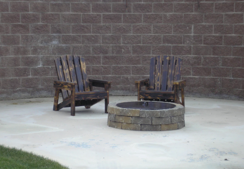 Two wooden chairs beside a circular stone fire pit on a concrete patio against a brick wall.