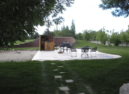 A serene outdoor patio with tables and chairs, surrounded by greenery and a gravel path leading to it.
