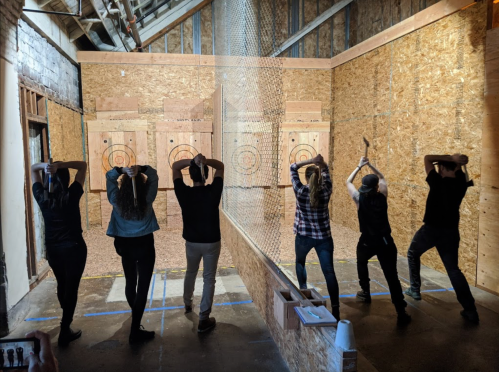 A group of six people stands in an indoor axe-throwing venue, preparing to throw axes at targets.