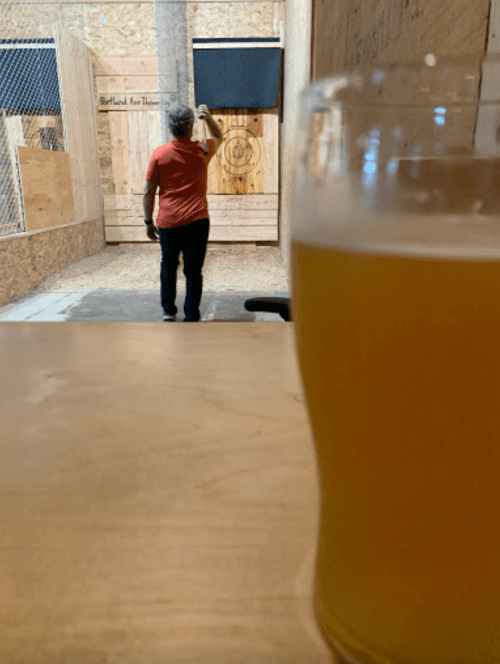 A person stands in an axe-throwing venue, with a glass of beer in the foreground.