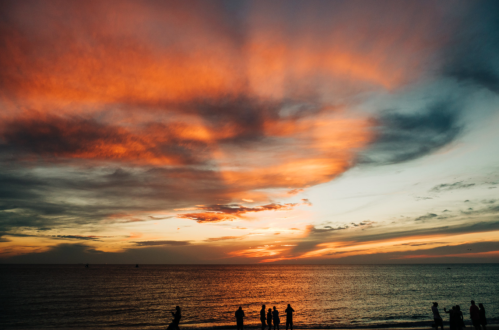 Silhouettes of people against a vibrant sunset over the ocean, with colorful clouds reflecting in the sky.
