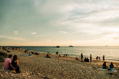 A beach scene at sunset with people walking, sitting, and enjoying the view by the water.