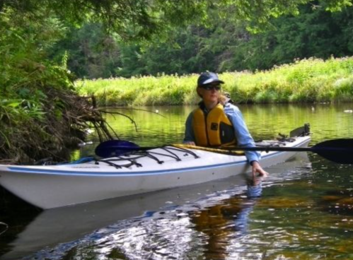 A person in a kayak on a calm river, surrounded by lush greenery and sunlight filtering through the trees.