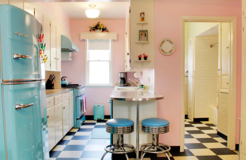 A retro-style kitchen with a pastel pink wall, blue fridge, checkered floor, and a small dining area with stools.
