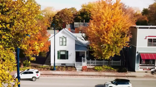A white house with a picket fence surrounded by vibrant autumn trees and a street with passing cars.
