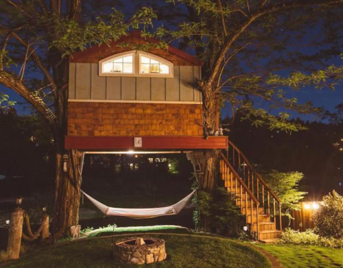 A cozy treehouse at night, illuminated with warm lights, featuring a hammock below and surrounded by greenery.