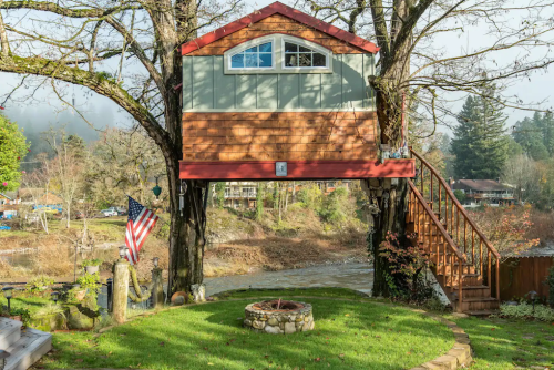 A treehouse with a red roof and large windows, nestled between trees by a river, with a fire pit and American flag nearby.