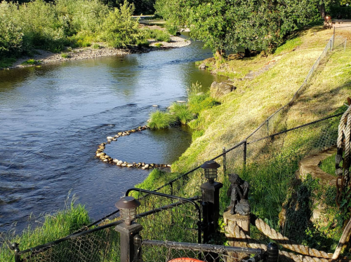 A serene river scene with a stone circle in the water, surrounded by lush greenery and a fence.