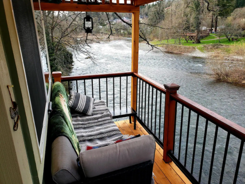 A cozy porch with a sofa overlooks a serene river surrounded by trees and greenery.