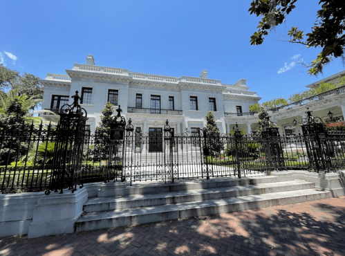 A grand mansion with a white facade, surrounded by a black wrought-iron fence and lush greenery under a clear blue sky.