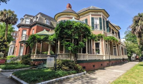 Historic Victorian-style house with a wraparound porch, surrounded by lush greenery and palm trees.