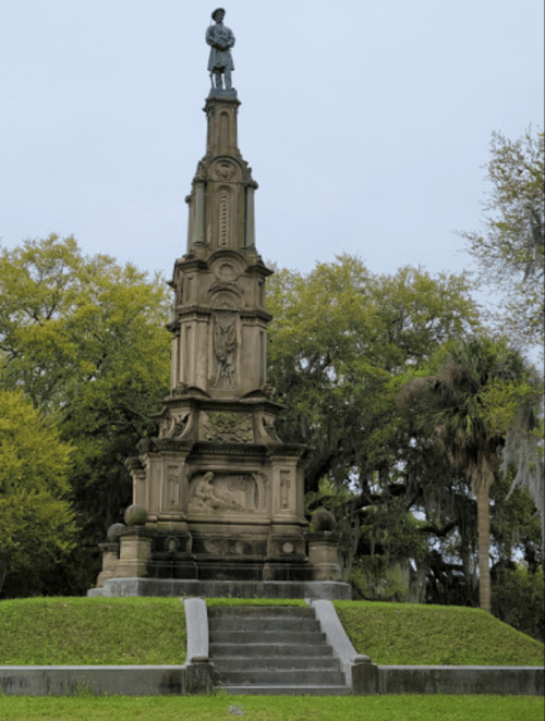 A tall stone monument with a statue on top, surrounded by greenery and trees under a cloudy sky.
