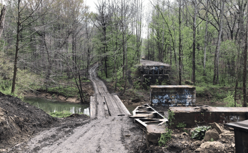 A muddy path leads to a creek, with remnants of a bridge and trees in early spring foliage surrounding the area.