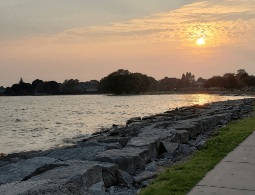Sunset over a calm lake, with rocky shoreline and trees in the background, casting a warm glow on the water.