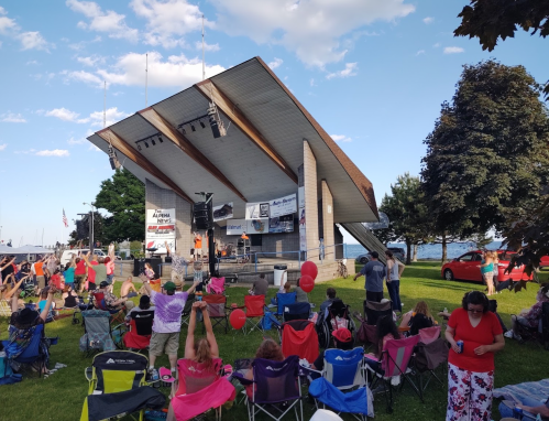 A crowd enjoys a live performance at an outdoor stage by the water, with colorful chairs and trees in the background.