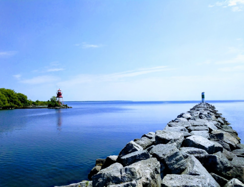 A rocky pier extends into calm blue waters, with two lighthouses visible in the distance under a clear sky.