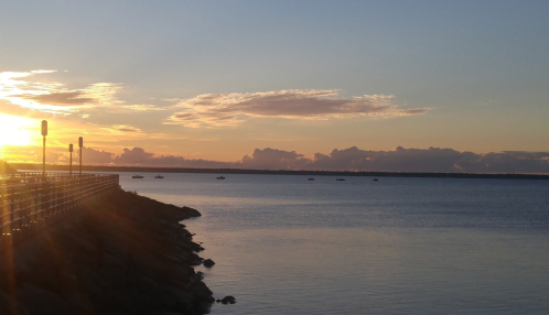 Sunset over calm waters, with a pier on the left and boats in the distance against a colorful sky.