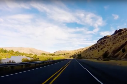 A scenic highway stretches through rolling hills under a blue sky with scattered clouds. Greenery lines the road.