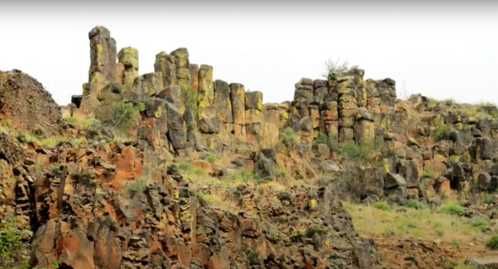 Rocky cliffside featuring tall, columnar formations and sparse vegetation under a cloudy sky.