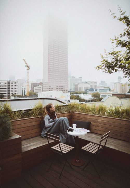 A woman sits wrapped in a blanket on a rooftop, overlooking a foggy cityscape with a tall building in the background.