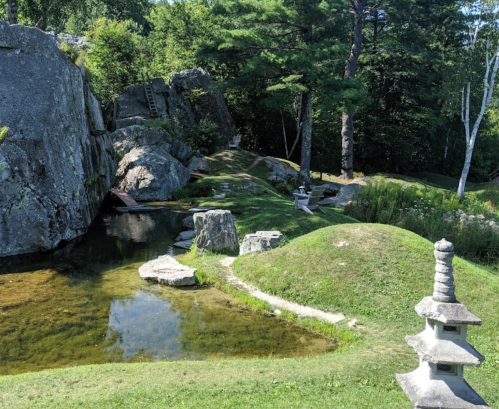 A serene landscape featuring a pond, rocky formations, and a stone lantern surrounded by lush greenery.