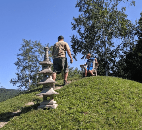 A man walks up a grassy hill towards a child sitting on a chair, with a stone pagoda structure nearby.