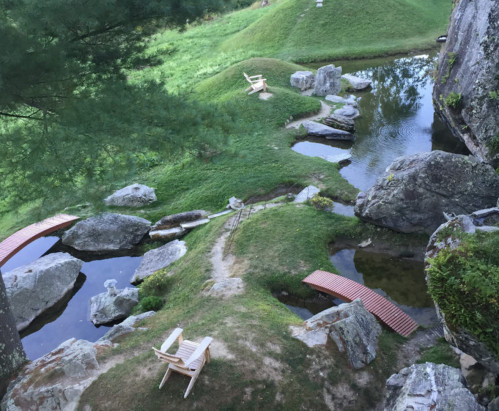 A serene landscape featuring a stream, rocks, wooden bridges, and chairs on grassy banks.