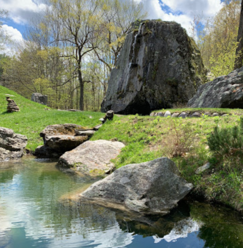 A serene landscape featuring a pond, large rocks, and lush greenery under a partly cloudy sky.