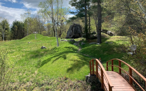 A wooden bridge crosses a grassy area with trees and rocks under a partly cloudy sky.