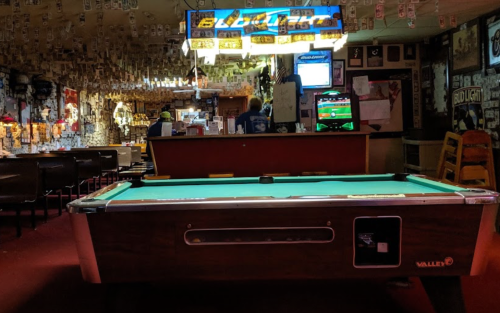A pool table in a dimly lit bar with vintage decor and a bartender in the background.