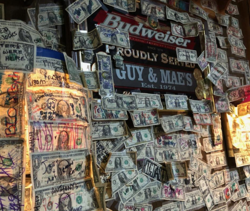 A wall covered in dollar bills with various signatures and messages, alongside a Budweiser sign.