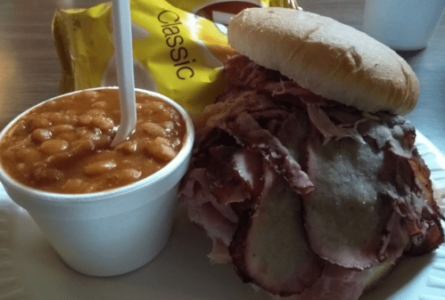 A plate with a large sandwich filled with sliced meat, a cup of baked beans, and a bag of chips in the background.