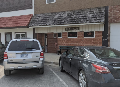 Exterior of G-M Tavern with two parked cars in front, featuring a brick facade and a wooden awning.