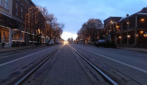 A quiet street at dusk, lined with trees and buildings adorned with festive lights, leading to a bright horizon.