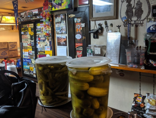 Two large jars of pickles sit on a bar counter, surrounded by various stickers and memorabilia on the wall behind.