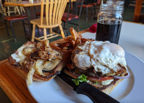 A burger topped with a fried egg and caramelized onions, served with fries and a dark beverage in a cozy restaurant setting.