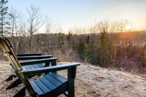 Three black Adirondack chairs overlook a serene landscape at sunset, surrounded by bare trees.