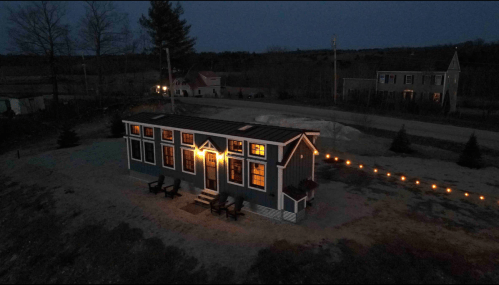 A cozy tiny house illuminated at night, surrounded by trees and string lights, with a gravel path leading to it.