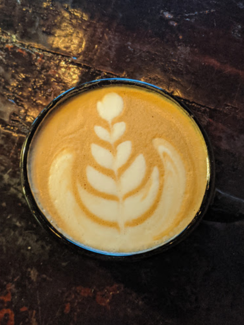 A close-up of a latte in a black cup, featuring a leaf-shaped foam art design on top.