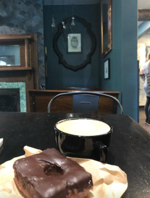 A chocolate-covered pastry on a paper napkin next to a black coffee cup, with a cozy café background.