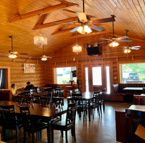 Cozy wooden restaurant interior with tables, ceiling fans, and large windows, featuring two diners at a table.