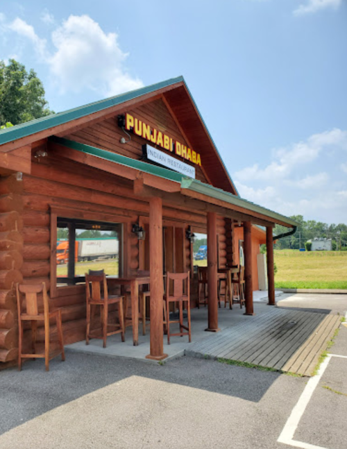 A wooden restaurant building with a sign reading "Punjabi Dhaba" and outdoor seating, set against a clear blue sky.
