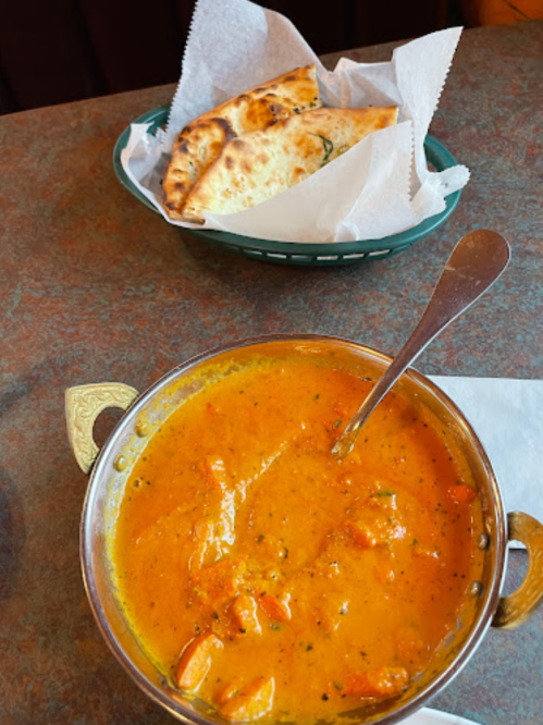 A bowl of creamy orange curry with vegetables and a spoon, alongside a basket of naan bread.