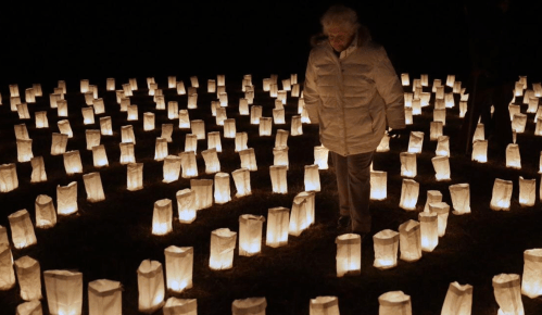 An elderly woman walks through a dark field illuminated by glowing lanterns arranged in a spiral pattern.