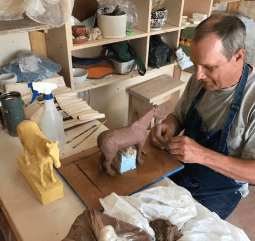 A man works on a clay horse sculpture in a studio, with tools and materials scattered around him.