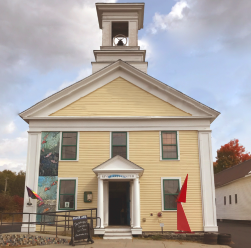 A yellow building with a bell tower, featuring colorful murals and red accents, houses the River Arts Center.