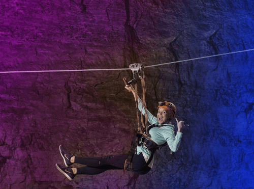 A person zip-lining against a colorful rock wall, smiling and making a playful gesture.