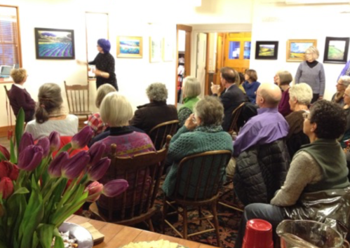 A group of people seated in a gallery, attentively watching a presentation with artwork displayed on the walls.
