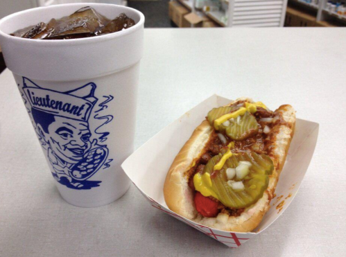 A hot dog topped with chili, pickles, mustard, and onions, next to a cup of iced drink on a table.